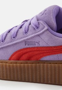 Fialový semišový teniska s tmavě červeným koženým pruhem, zaoblenou špičkou a texturovanou gumovou podrážkou. Má stahovací tkaničky ve stejném odstínu a vyražené logo.