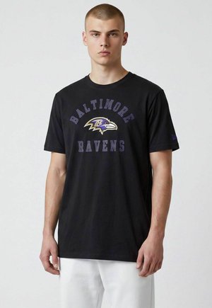 Jonge man met kort haar die een zwart Baltimore Ravens T-shirt en een witte broek draagt, staand tegen een effen lichte achtergrond.