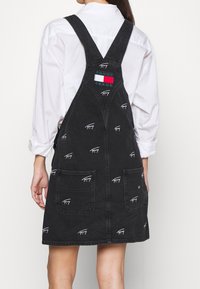 Kvinna som bär en svart denimklänning med broderade vita logotyper och en märkespåse, över en vit långärmad skjorta, sedd bakifrån.