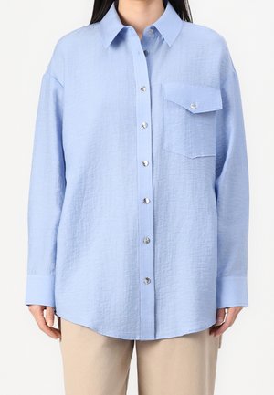 Femme portant une chemise bleu clair texturée à boutons avec une poche poitrine unique, associée à un pantalon beige, vue du cou aux hanches.
