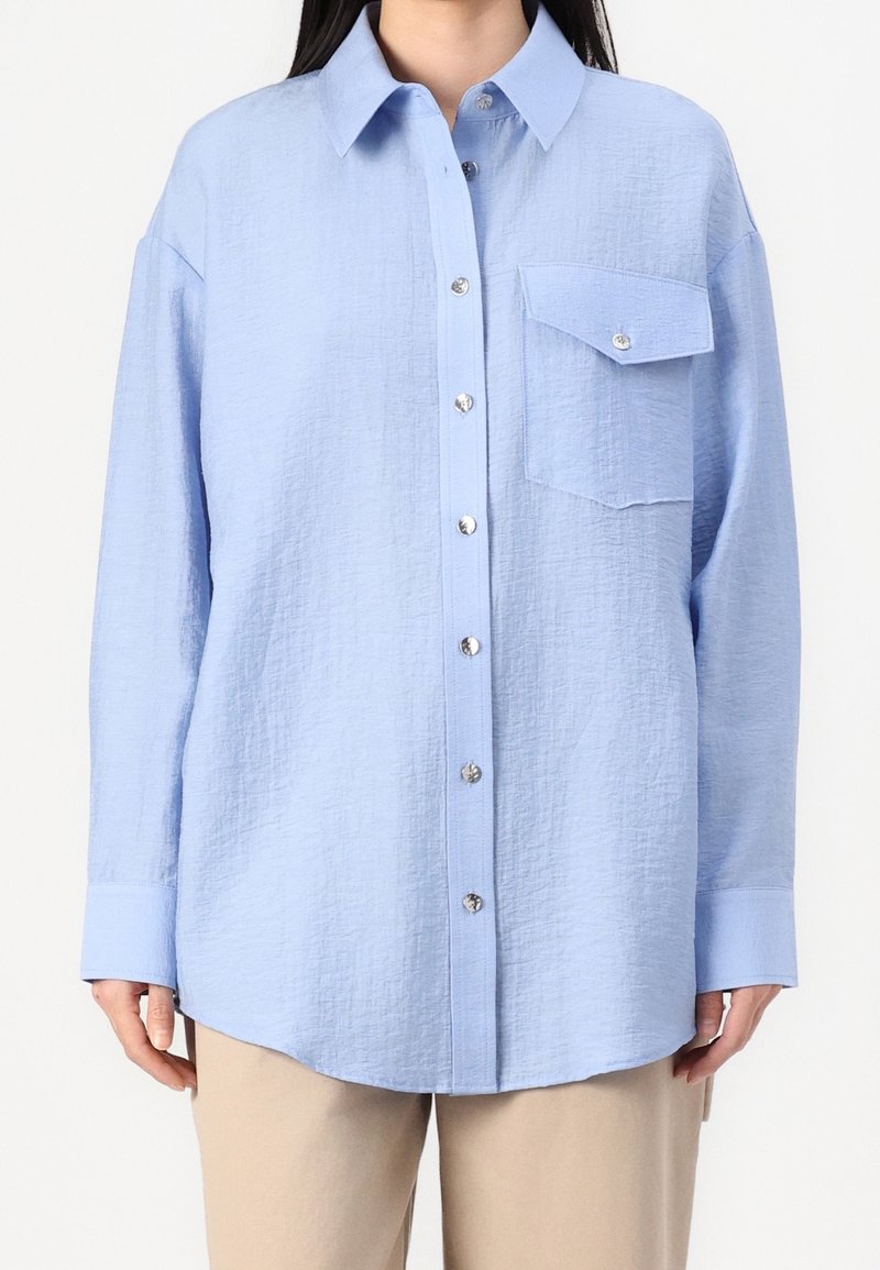 Femme portant une chemise bleu clair texturée à boutons avec une poche poitrine unique, associée à un pantalon beige, vue du cou aux hanches.