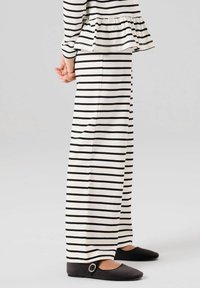 Personne portant un pantalon à rayures horizontales noires et blanches assorti à un haut, debout les mains jointes derrière le dos, portant des ballerines noires à boucle visibles.