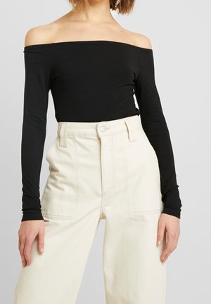 Vrouw draagt een zwarte off-shoulder top met lange mouwen en een crème broek met hoge taille en wijde pijpen, voorzien van voorzakken met klep.