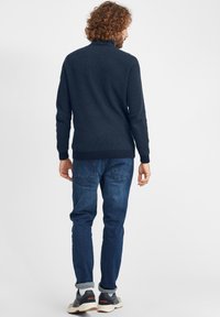 Mornarsko pleteno pulover z visokim ovratnikom, rebrastimi manšeti in robom, v kombinaciji z modrimi jeans hlačami. Vidna podrobnost žepa na hlačah in superge.