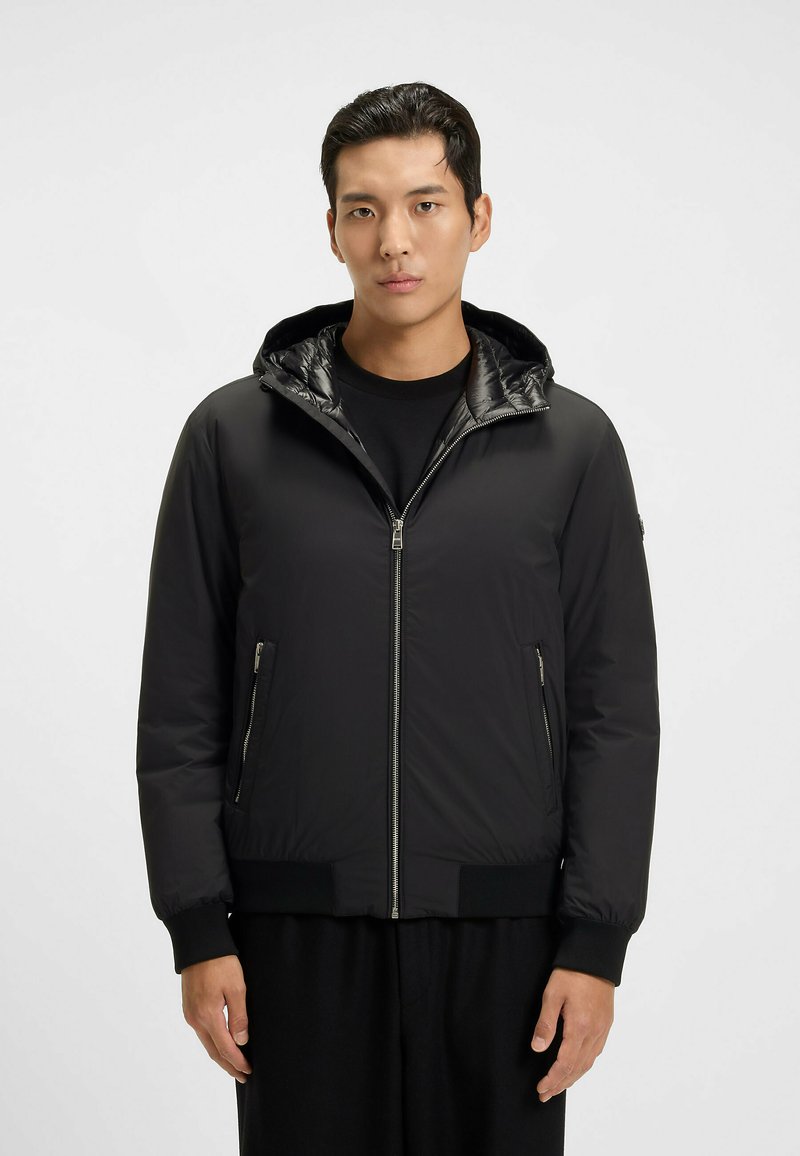 Ung man som bär en svart zip-up huvtröja med zip-fickor på sidorna, stående mot en plain ljus bakgrund.