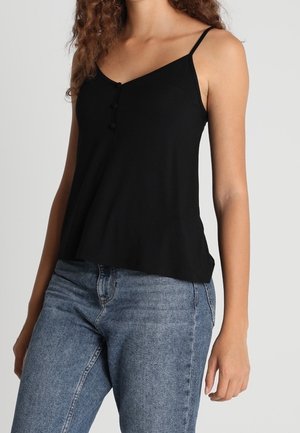 Kobieta ubrana w czarny, bezrękawnik z dekoltem w serek i guzikami, połączony z niebieskimi dżinsami na jednolitym tle.