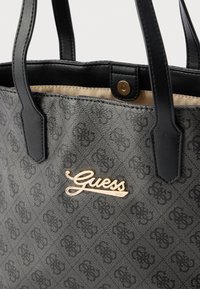 Čierna vzorovaná taška na rameno s koženými rukoväťami. Na prednej strane je zlaté logo „Guess“. Vnútro je béžové a obsahuje zapínanie na cvok.