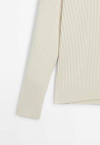 Pull en maille crème à manches longues avec un motif côtelé vertical et de larges poignets côtelés, sur un fond blanc uni.