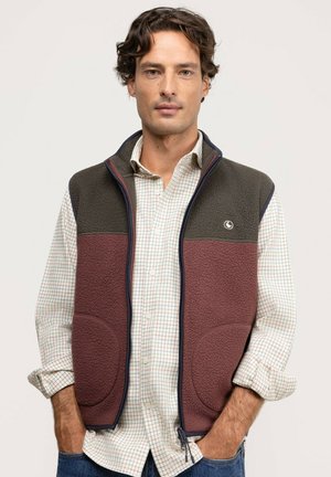 Hombre con camisa de cuadros con las mangas remangadas y un chaleco polar bicolor con las manos en los bolsillos, de pie frente a un fondo liso.