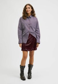 Chemise à manches longues à boutons avec rayures bleues et rouges, portée avec une jupe en sequins bordeaux. Bottes noires montantes jusqu'au genou avec des sangles et des boucles latérales.