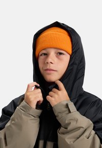 Jongen met een oranje gebreide muts en een beige-zwarte winterjas, die de rits van de jas bij de capuchon aanpast.