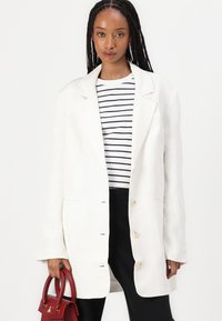 Kvinne med langt flettehår som har på seg en hvit oversized blazer, svart og hvitstripet skjorte, svarte bukser og holder en rød veske.