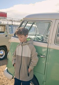 Leichte, zweifarbige Jacke in Beige und Grau, mit Kapuze und Reißverschluss. Das Kind lehnt sich an einen vintage grünen Van.