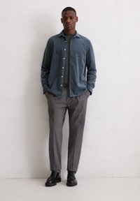 Mann in einem blauen Hemd mit Knöpfen, grauen Hosen und schwarzen Schnürschuhen, der mit den Händen in den Taschen vor einer schlichten Wand steht.