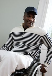 Gestreepte trui in marineblauw en crème met een ritskraag, gecombineerd met witte broek. Man zittend in een rolstoel, met een marineblauwe pet.