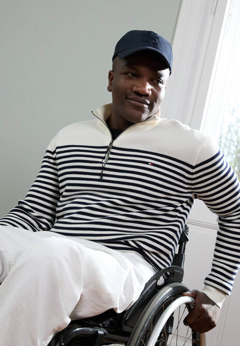 Gestreepte trui in marineblauw en crème met een ritskraag, gecombineerd met witte broek. Man zittend in een rolstoel, met een marineblauwe pet.