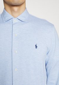 Vaaleansininen nappikauluspaita, jossa on rakenteellinen kuvio, ja vasemmassa rinnassa on pieni tummansiniharmaa brodeerattu logo. Napit ovat valkoiset.