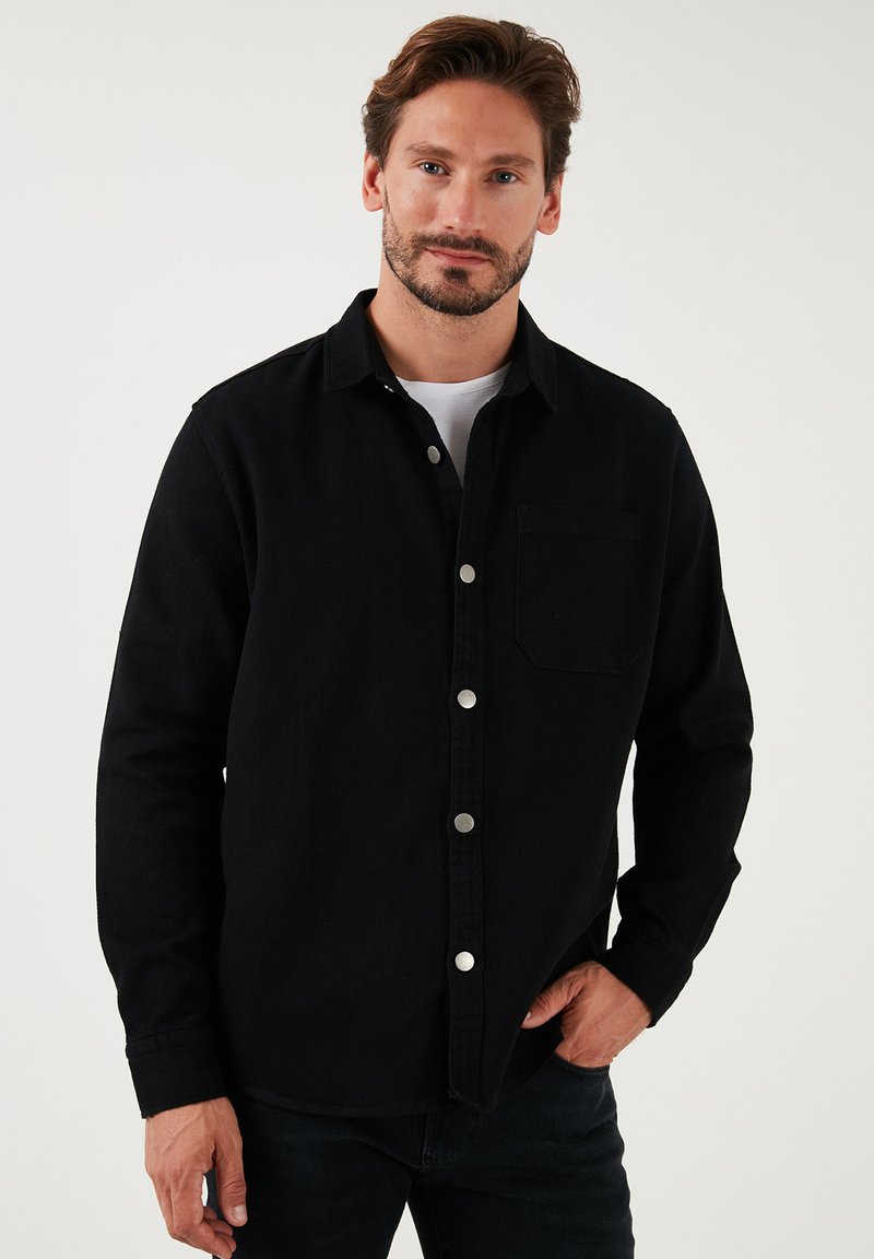 Camisa negra de botones con cuello, que presenta un único bolsillo en el pecho y botones a presión plateados. Fabricada en una tela texturizada con mangas largas.