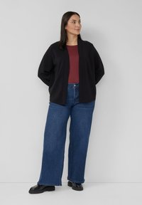 Vrouw staande met handen achter de rug, gekleed in een zwarte open jas, bordeauxrode top, wijde blauwe jeans en zwarte loafers, tegen een effen achtergrond.