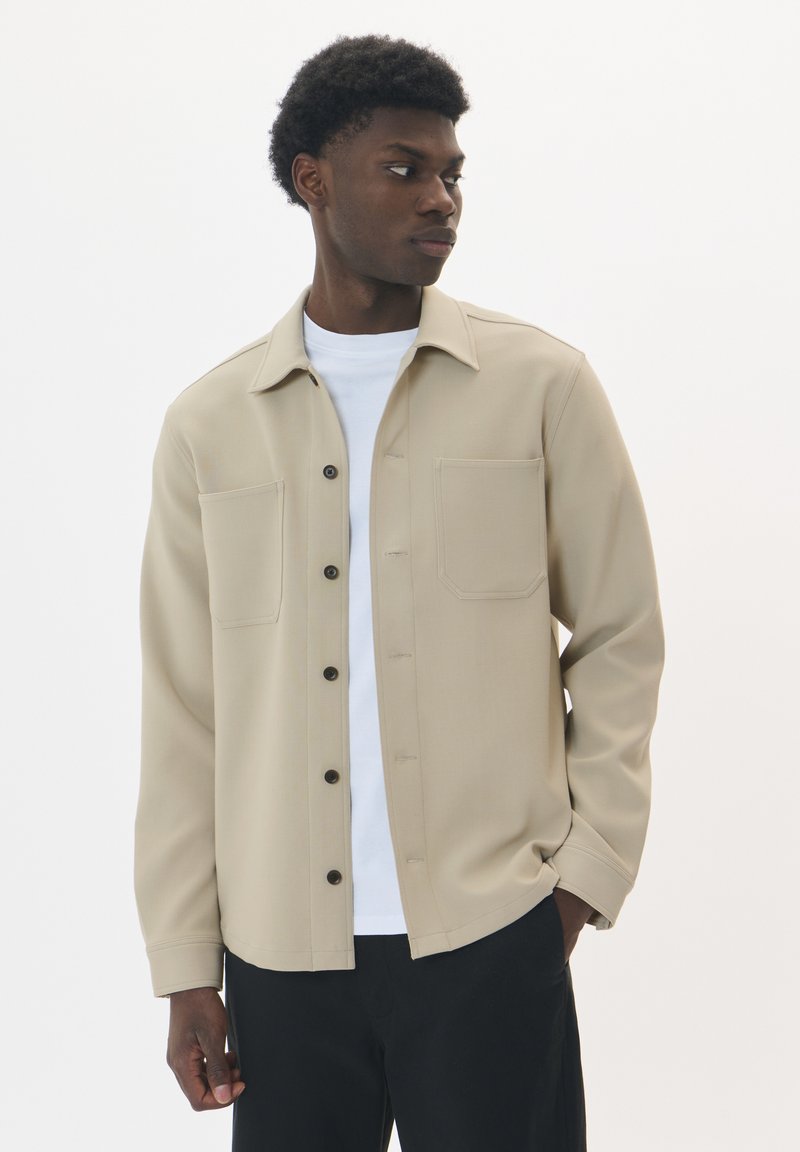 Giacca beige con bottoni, colletto, due tasche sul petto e bottoni scuri, indossata sopra una camicia bianca. Tessuto morbido con una texture liscia.