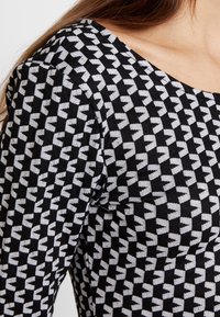 Czarny dzianinowy top z geometrycznym wzorem białych kształtów, o miękkiej teksturze i szerokim dekolcie. Wzór jest jednolity na całej tkaninie.