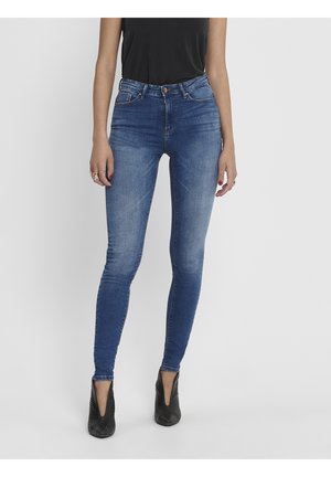Kvinna i högmidjade blå skinny jeans med urtvättad detaljering, svart ärmlös topp, svarta spetsiga skor och ringar på fingrarna.