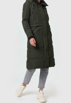 Personne portant un long manteau matelassé vert olive avec de grandes poches, des jeans gris coupés au-dessus des chevilles et des baskets blanches épaisses sur un fond uni.