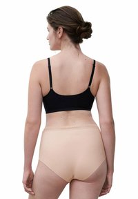 Frau mit zurückgebundenem Haar, die einen schwarzen BH und hautfarbene, hochgeschnittene Unterhosen trägt, stehend und mit dem Rücken zu einer weißen Hintergrund.
