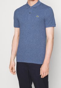 Blå polo skjorta i texturerat tyg, med klassisk krage, tvåknaps placket och en liten grön Lacoste-logotyp på bröstet.
