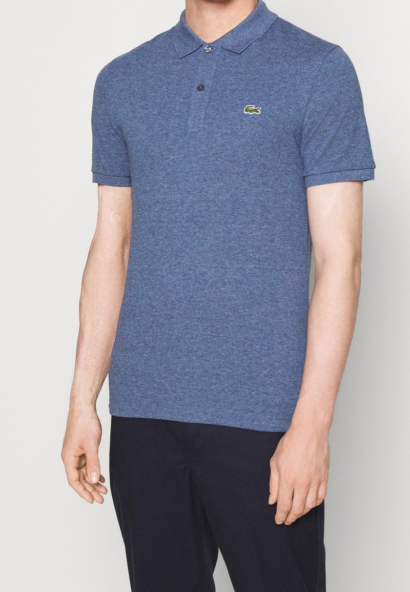 Blå polo skjorta i texturerat tyg, med klassisk krage, tvåknaps placket och en liten grön Lacoste-logotyp på bröstet.