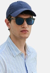 Gorra texturizada azul marino, gafas de sol rectangulares negras con lentes oscuros, y camisa de botones a rayas azul claro con un cuello informal.