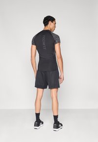 Εφαρμοστή μαύρη αθλητική μπλούζα με κοντά μανίκια και διακοσμητικές λεπτομέρειες από δίχτυ, συνδυασμένη με φαρδιά μαύρα σορτς με υφή και λευκές πλευρικές ρίγες.