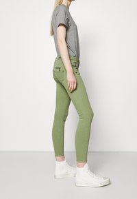 Green skinny jeans with zippered pockets and a mid-rise waist, paired with a gray t-shirt and white high-top sneakers.