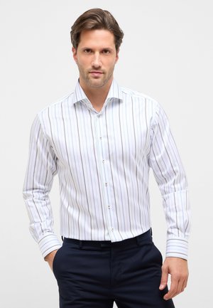 Uomo con capelli castani corti che indossa una camicia a righe verticali bianche, blu e grigie con bottoni e pantaloni blu scuro, in piedi con una mano in tasca.