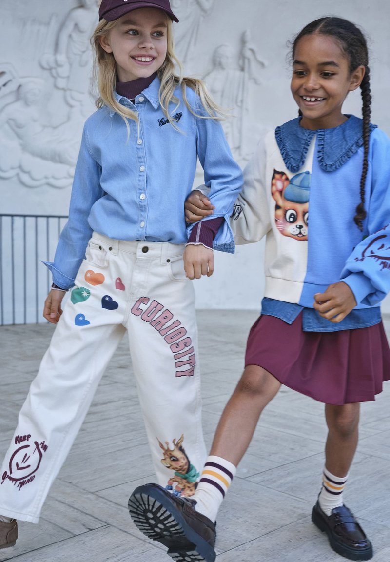 Deux filles marchent bras dessus bras dessous, l'une porte un pantalon blanc avec des patchs colorés et une chemise bleue, l'autre une jupe violette et un pull bleu.