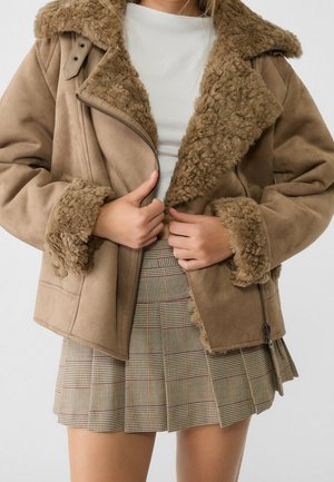 Persona con chaqueta de piel sintética marrón claro con cuello y puños de piel sobre una camiseta blanca y una falda plisada de cuadros, ajustando la cremallera de la chaqueta.