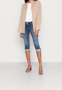 Femme portant un t-shirt blanc, une chemise oversize beige à rayures avec boutons, un jean court retroussé et des sandales à talons marron avec des lanières.