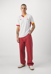Maillot de football blanc avec des accents rouges et orange, comprenant un col en V, des manches courtes et un logo. Associé à un jean rouge et des baskets blanches.