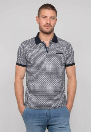 Jeune homme aux cheveux courts et à la barbe, portant un polo à motifs bleu marine et blanc et un jean bleu clair, les mains dans les poches, fond neutre.