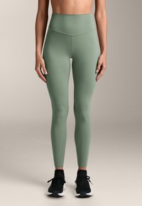 Visoko strukirane tajice u prigušenoj zelenoj boji, izrađene od rastezljivog materijala, s glatkom teksturom i bez vidljivih šavova, u kombinaciji s crnim tenisicama.