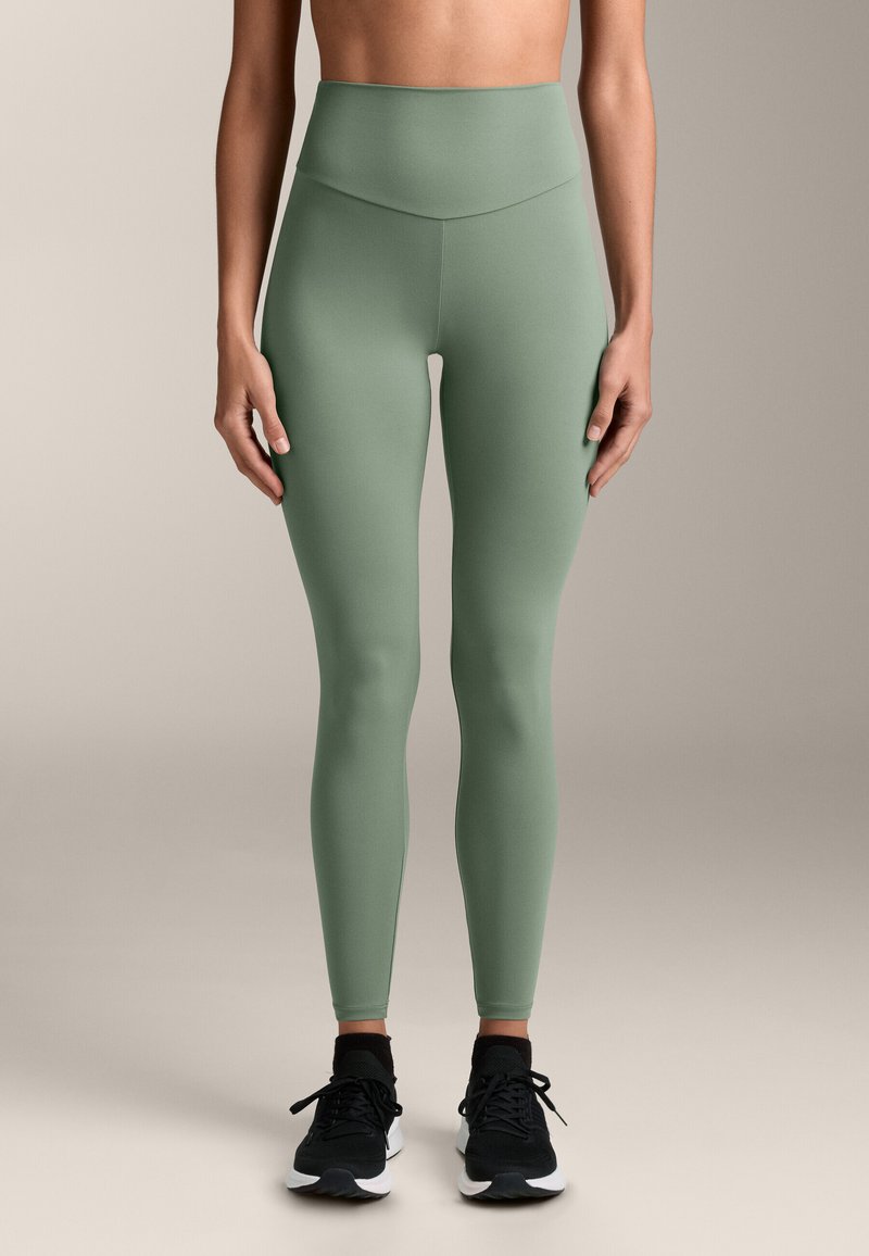 Visoko strukirane tajice u prigušenoj zelenoj boji, izrađene od rastezljivog materijala, s glatkom teksturom i bez vidljivih šavova, u kombinaciji s crnim tenisicama.