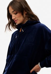 Granatowy welurowy hoodie z wysokim kołnierzem, ściągaczem i przednimi kieszeniami. Wyposażony w haftowane logo i paski na mankietach. Miękka tekstura.