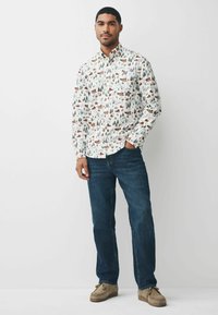 Langærmet skjorte med et vinterlandskabsmønster med huse og træer i nuancer af grøn og brun på en hvid baggrund. Båret med blå jeans.