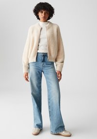 Frau steht in einer cremfarbenen, flauschigen Zip-Jacke, einem weißen Rollkragenpullover, hellblauen weit geschnittenen Jeans und beigen Turnschuhen vor einem grauen Hintergrund.