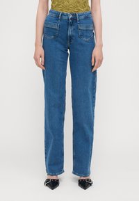 Jeans de ganga azul de cintura alta com corte reto, cinco bolsos e um desbotamento subtil. Combinados com sapatos pretos de bico fino.