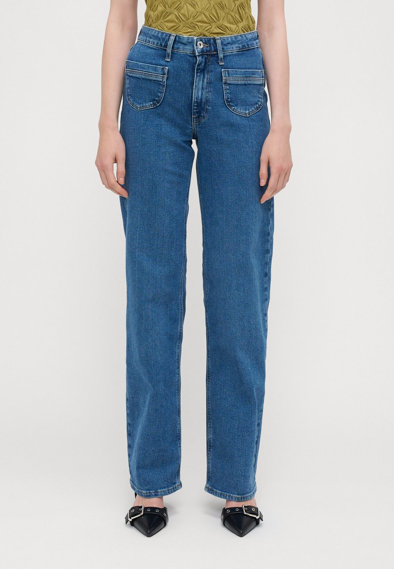 Jeans de ganga azul de cintura alta com corte reto, cinco bolsos e um desbotamento subtil. Combinados com sapatos pretos de bico fino.