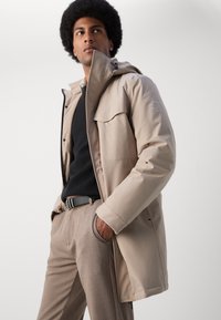 Chaqueta de exterior beige con capucha, diseñada con una textura suave y bolsillos frontales, combinada con un suéter oscuro y pantalones marrones.