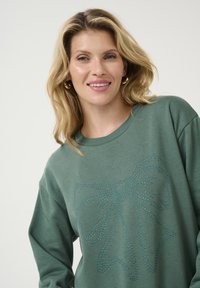 Sweatshirt vert en tissu doux, présentant un grand nœud en strass scintillant sur le devant. Coupe décontractée avec un col rond.