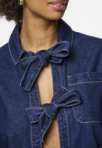 Närbild av en person som bär en mörkblå denimjacka med två flugor som säkrar framdelen.