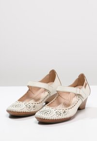 Valkoiset nahkaiset Mary Jane -kengät, joissa on leikattu kukkakuvio, säädettävä hihna ja matala puinen korko; sisältää myös ommellun pohjan yksityiskohdan.